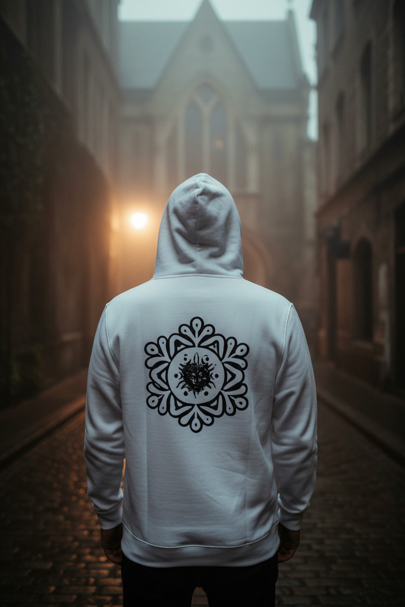 A person viewed from behind wearing a white hoodie with a large black mandala-style graphic on the back, standing in a narrow cobblestone alley with a blurred building and warm light in the distance.