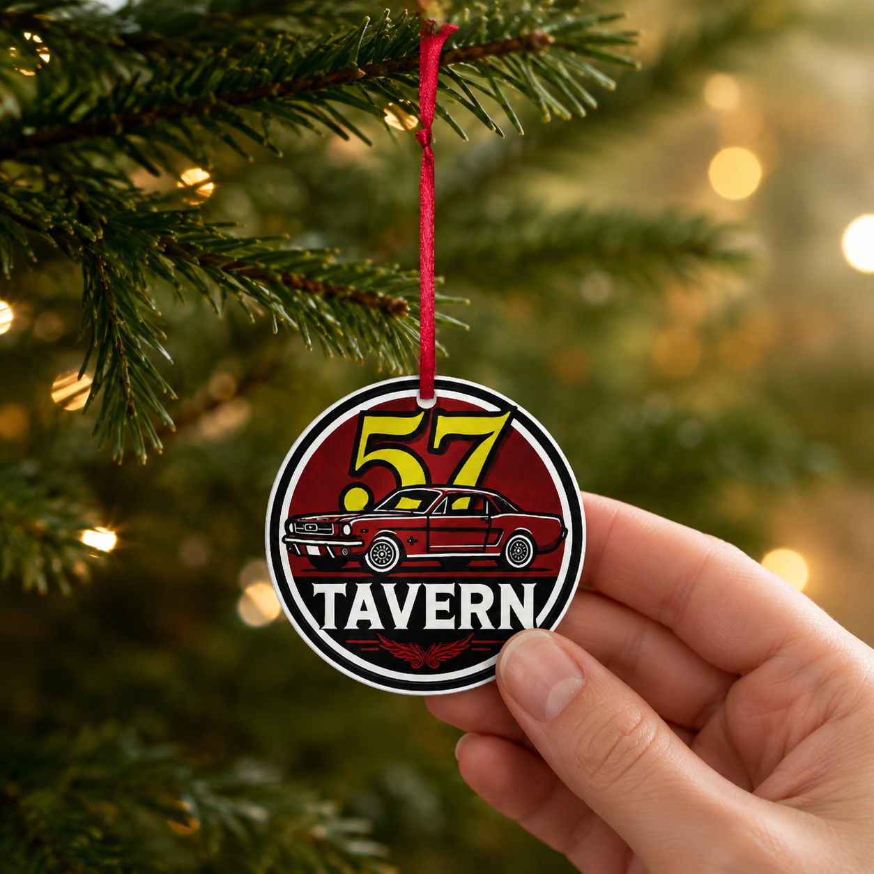 A hand holds a round Christmas ornament featuring a red car and the words “57 TAVERN” against a blurred Christmas tree background with warm lights.