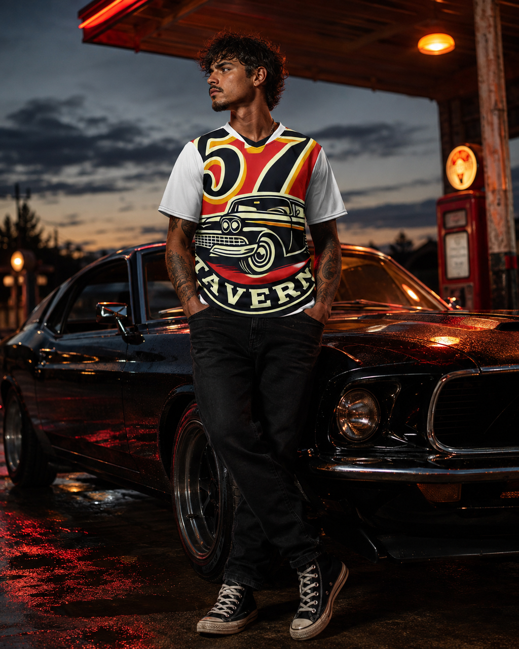 A man stands in front of a black classic muscle car at a gas station at dusk, wearing a white t-shirt with a large vintage car graphic and the word 'MAVERICK' in red, yellow, and black.