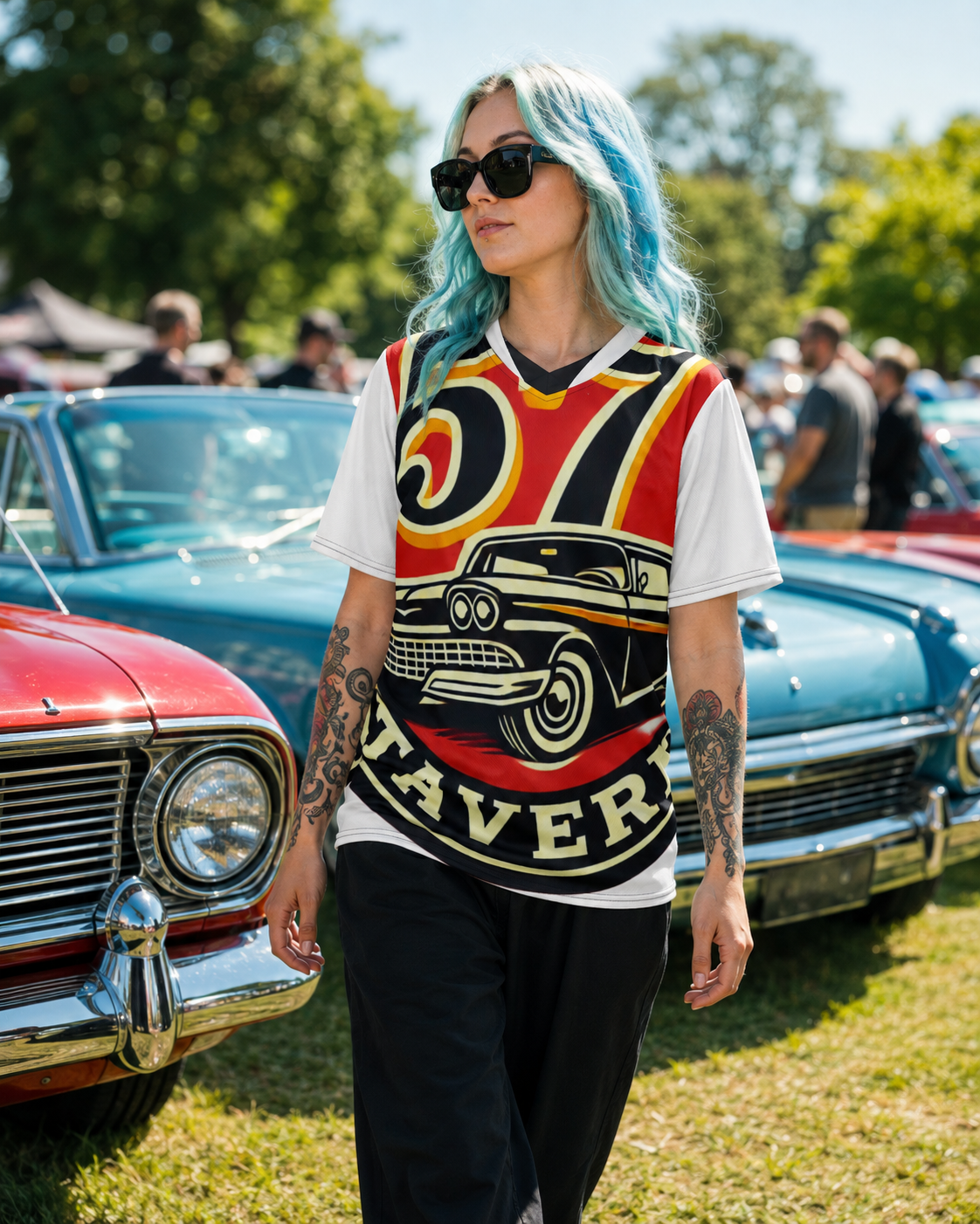 A person with bright blue hair and sunglasses stands outdoors at a car show wearing a white graphic t-shirt with a red, yellow, and black vintage-style print and the word 'MAVERICK' visible, paired with black pants.