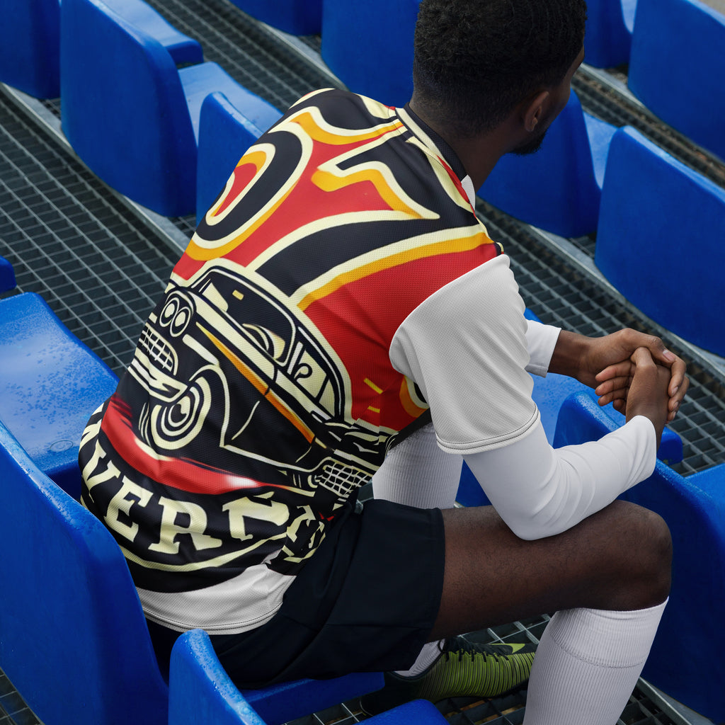 A man sits on blue stadium seats wearing a black, red, and yellow graphic jersey over a white long-sleeve shirt and black shorts.