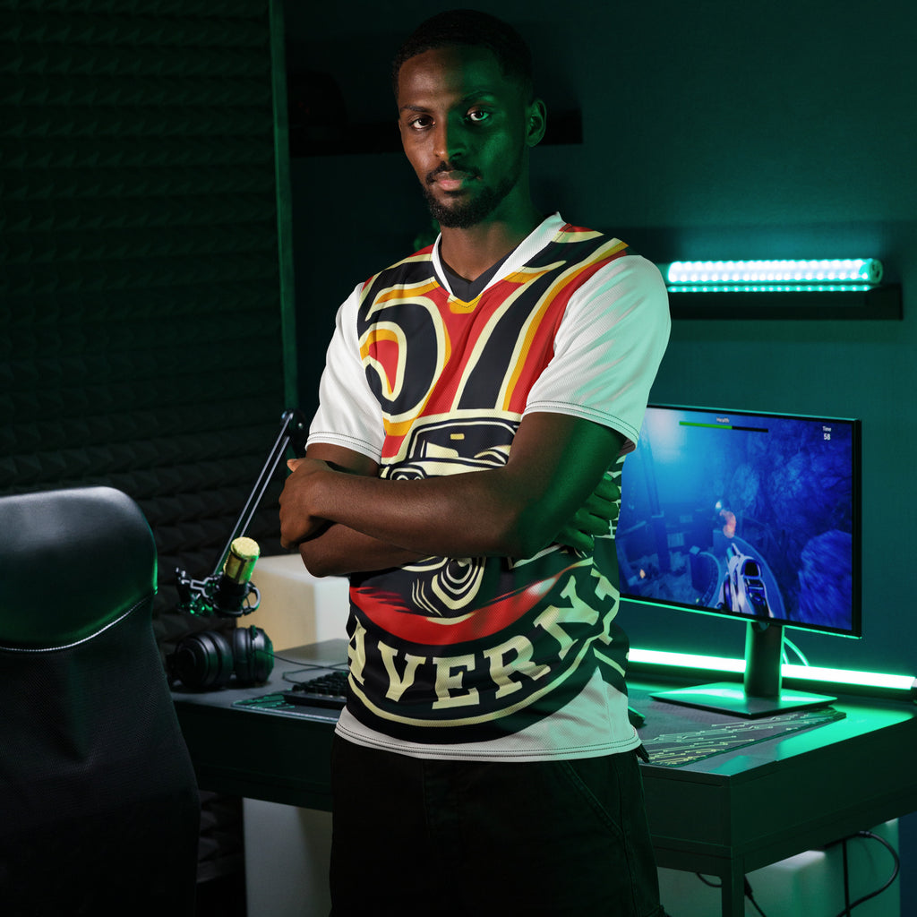 A man stands in a gaming room wearing a white t-shirt with a bold black, red, and yellow graphic design. The room features a desk with a computer monitor, a microphone, and green LED lighting.