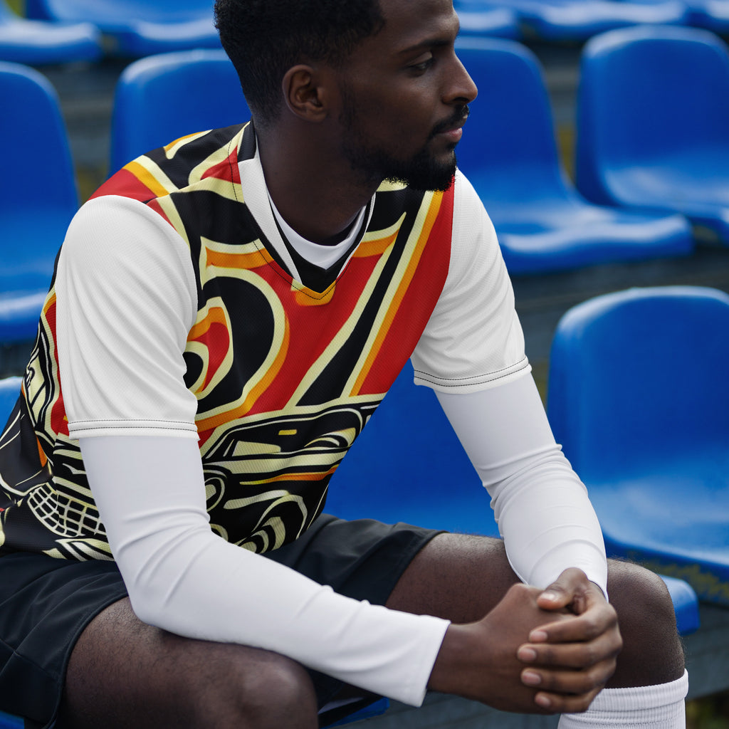 A man sitting in a stadium wearing a long-sleeve athletic jersey with a bold red, black, yellow, and white graphic pattern over a white long-sleeve base, with blue stadium seats in the background.