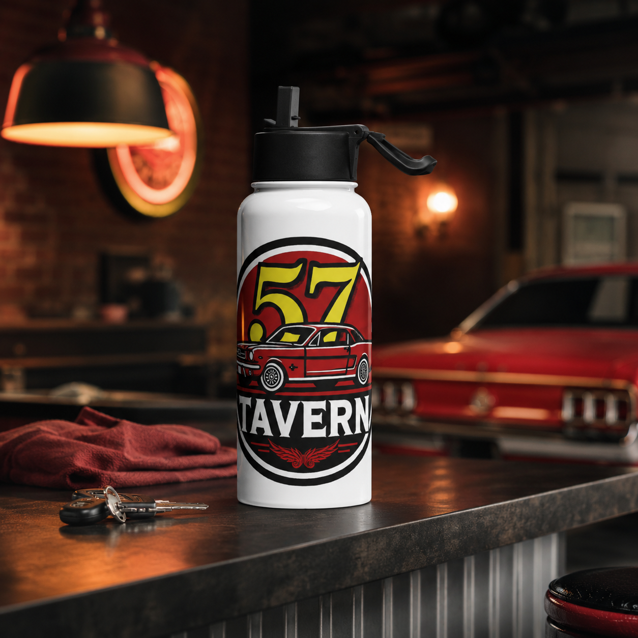 A white insulated water bottle with a black lid stands on a dark counter in a garage-like setting. The bottle features a circular red, yellow, black, and white graphic with the text '57 TAVERN' and an illustration of a car.