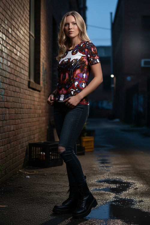 A woman stands in a wet urban alleyway wearing a short-sleeve crewneck t-shirt with a vibrant, multicolored graphic print, paired with dark ripped jeans and black combat boots.
