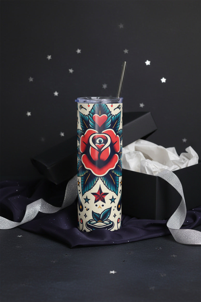 A tall insulated tumbler with a metal straw, featuring a traditional tattoo-style rose and star pattern in red, teal, black, and cream. A clear plastic lid is shown beside the tumbler on a solid white background.