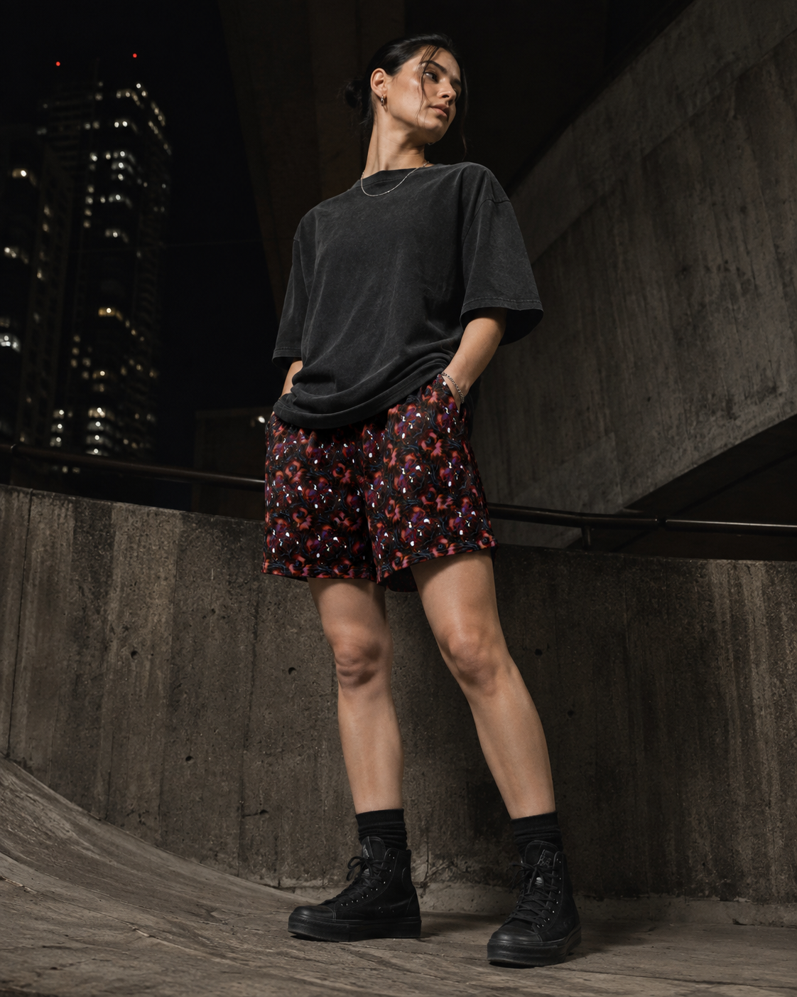 A model stands in a concrete urban setting at night, wearing a dark charcoal oversized t-shirt, red shorts with a black and white floral pattern, and black lace-up boots.