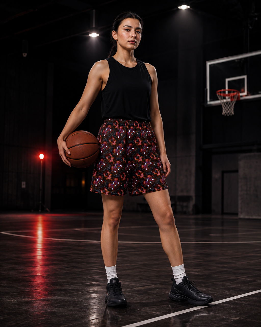 A woman stands on an indoor basketball court holding a basketball, wearing a black sleeveless top and black shorts with a colorful floral pattern, white socks, and black sneakers.
