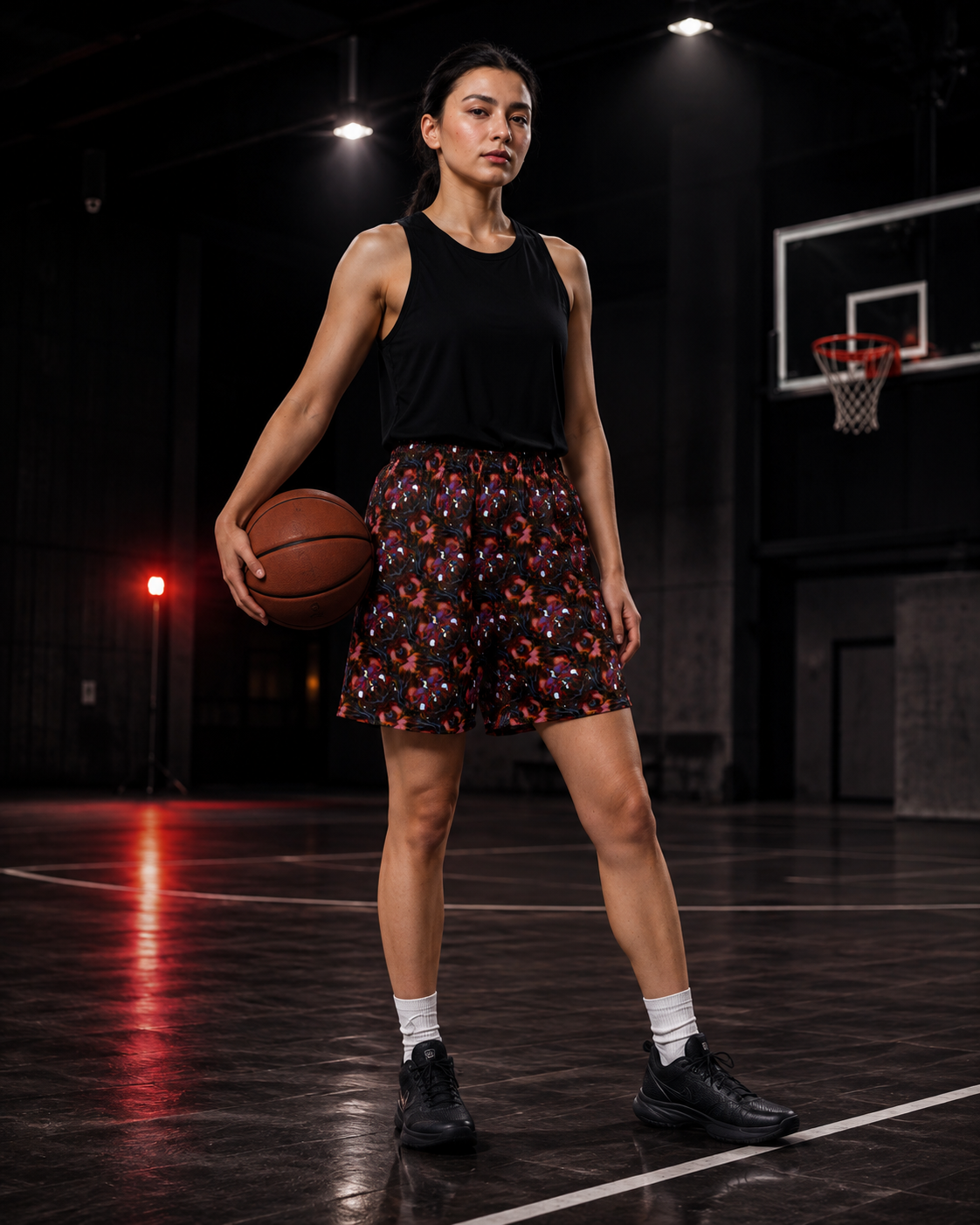 A woman stands on an indoor basketball court holding a basketball, wearing a black sleeveless top and black shorts with a colorful floral pattern, white socks, and black sneakers.