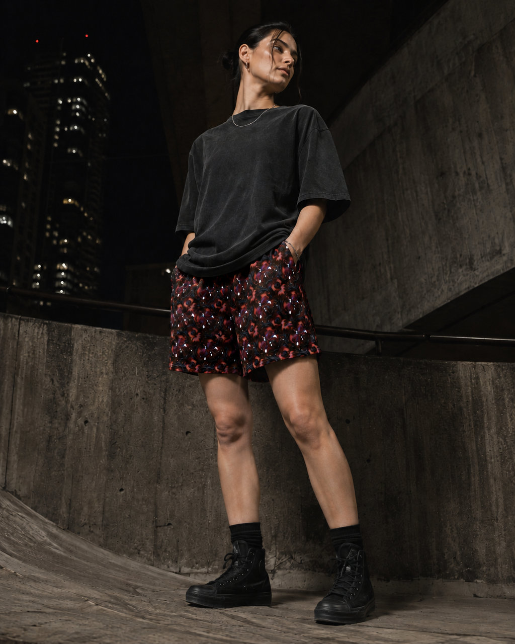 A model stands in a concrete urban setting at night, wearing a black oversized t-shirt, red shorts with a small floral pattern, and black combat boots.