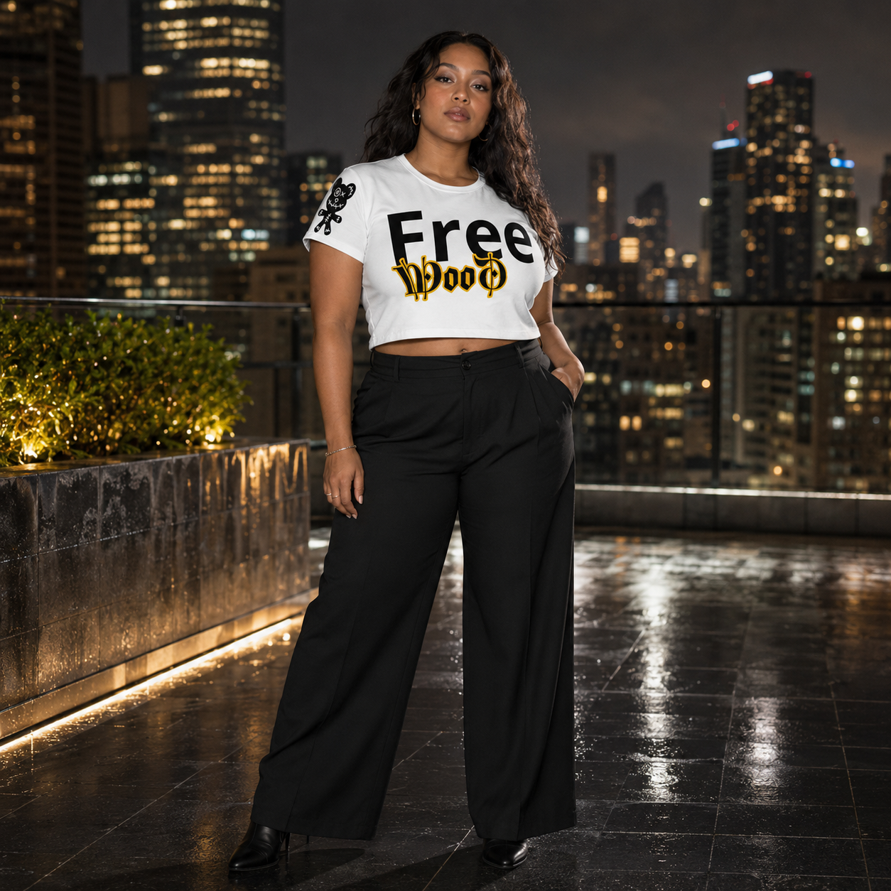A woman stands on a rooftop at night wearing a white graphic crop top with 'Free Wood' text and black wide-leg pants, with a city skyline in the background.