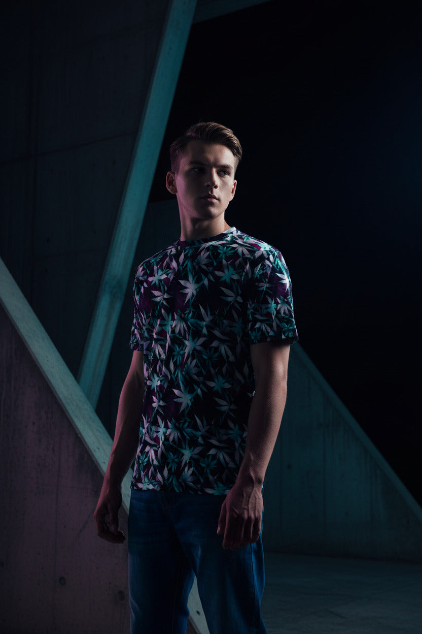A man wearing a short-sleeve crewneck t-shirt with an all-over cannabis leaf pattern in purple, white, and teal, paired with blue jeans against a plain white background.