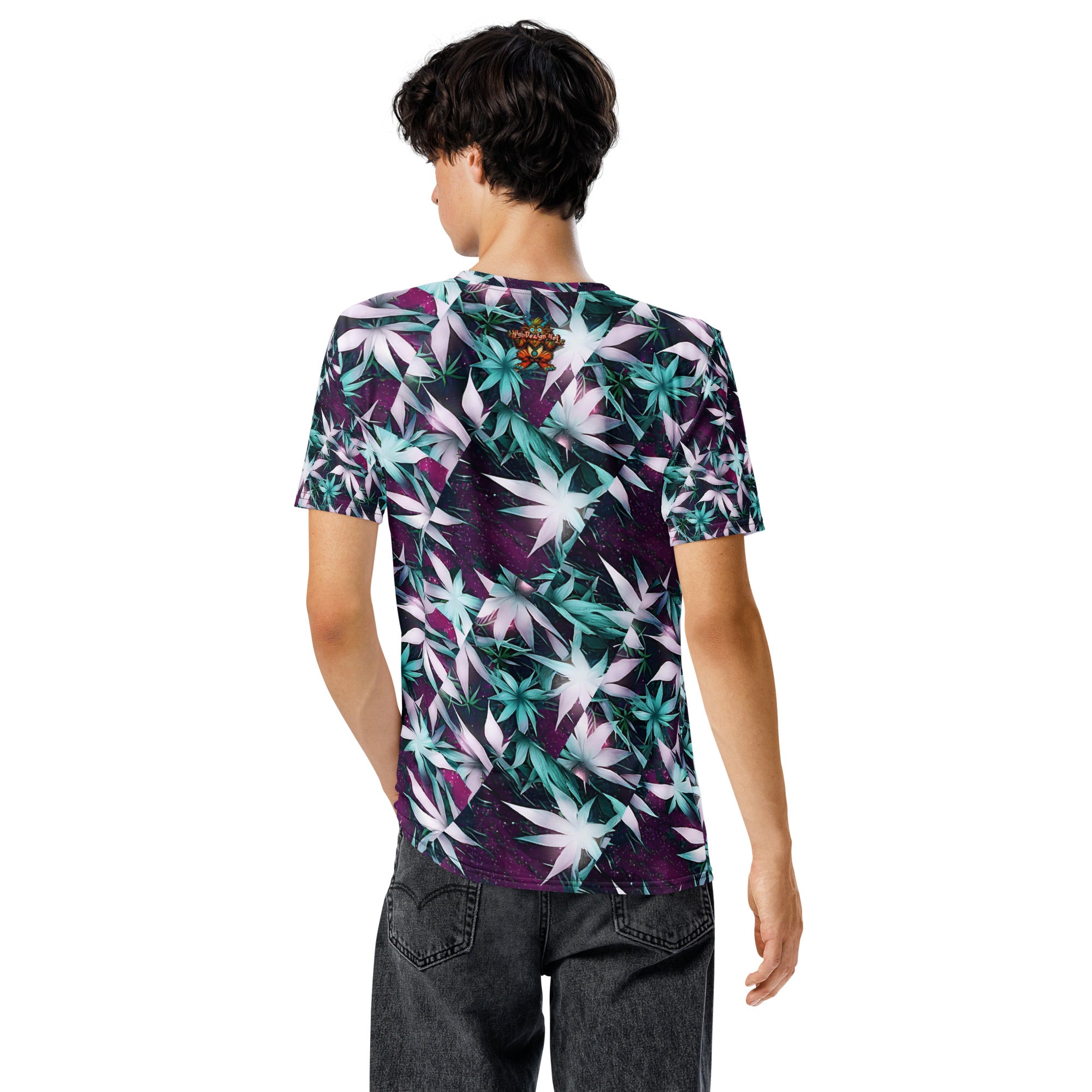 A man viewed from the back wearing a short-sleeve crewneck t-shirt with a vibrant purple, teal, and white leaf pattern, paired with dark gray jeans, against a plain white background.