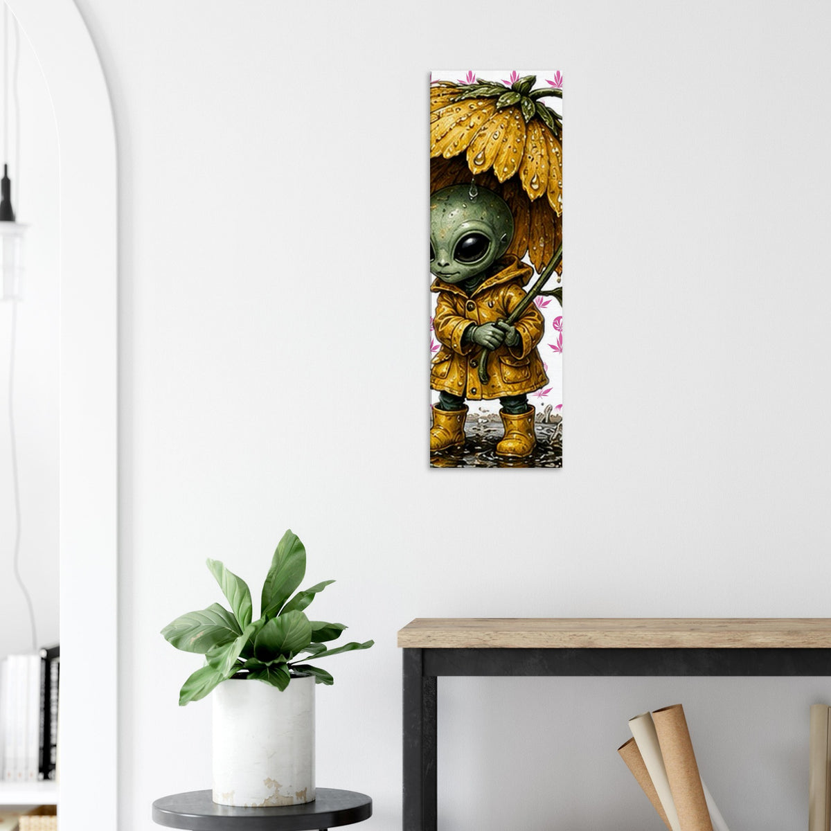 A vertical wall art print featuring a cute green alien in a yellow raincoat under a flower-shaped umbrella, displayed on a white wall above a console table with a potted plant.