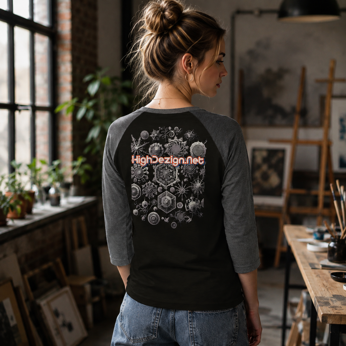 A woman stands in an art studio wearing a black and gray raglan baseball t-shirt with a detailed white mandala graphic on the back and the text 'HighDesign.Net' in orange.