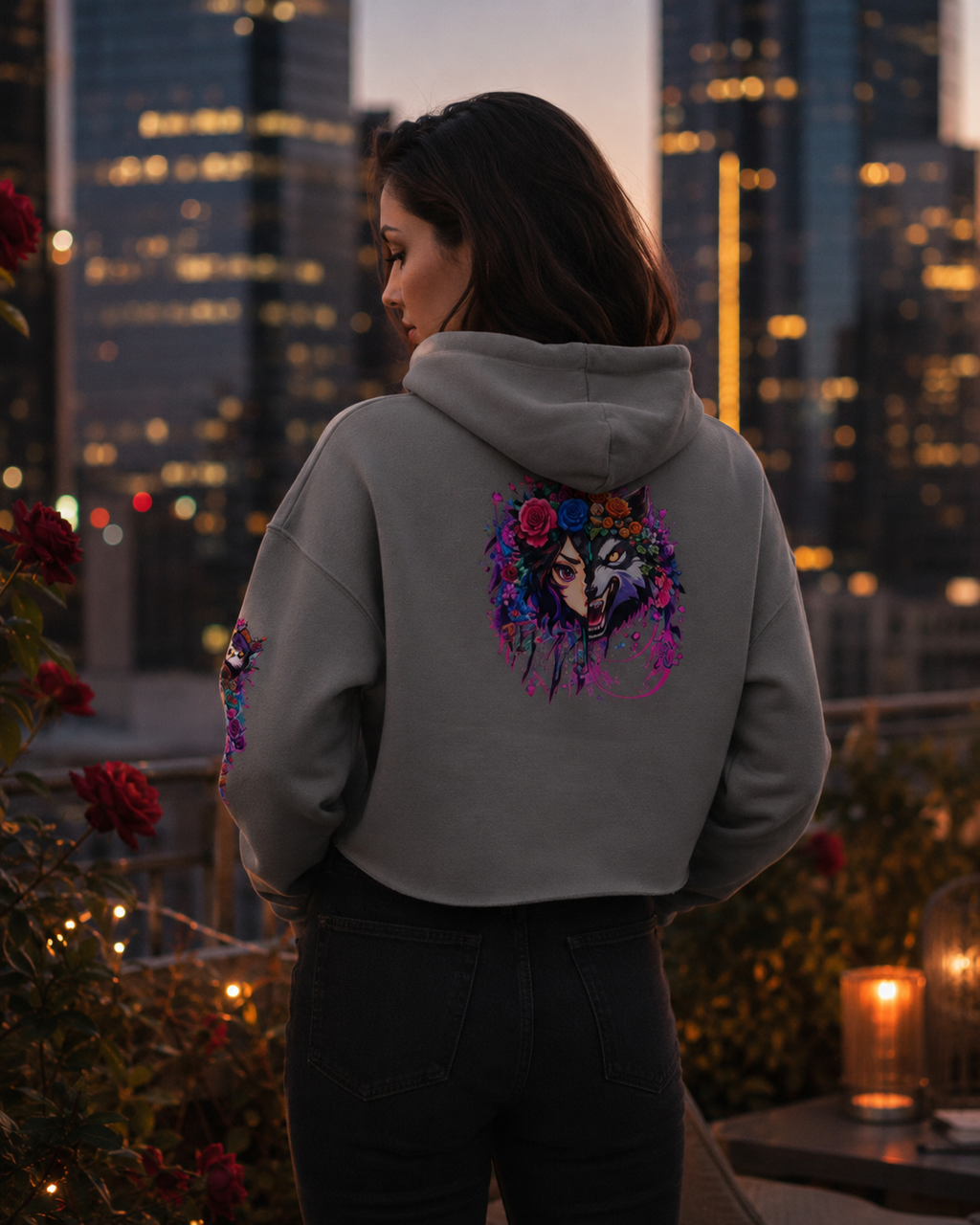 A woman stands on a rooftop at dusk wearing a gray cropped hoodie with a colorful floral and skull graphic on the back, paired with black pants. City buildings and warm lights are visible in the background.