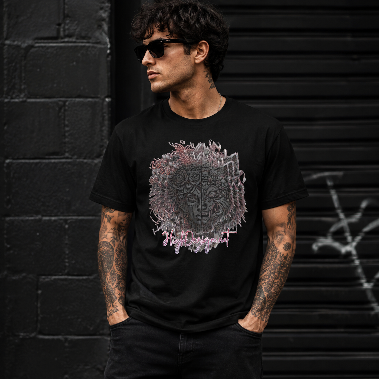 A man wearing a black graphic t-shirt with a detailed skull design and the text 'Hells Angels' in pink, standing in front of a dark brick wall and metal shutter.