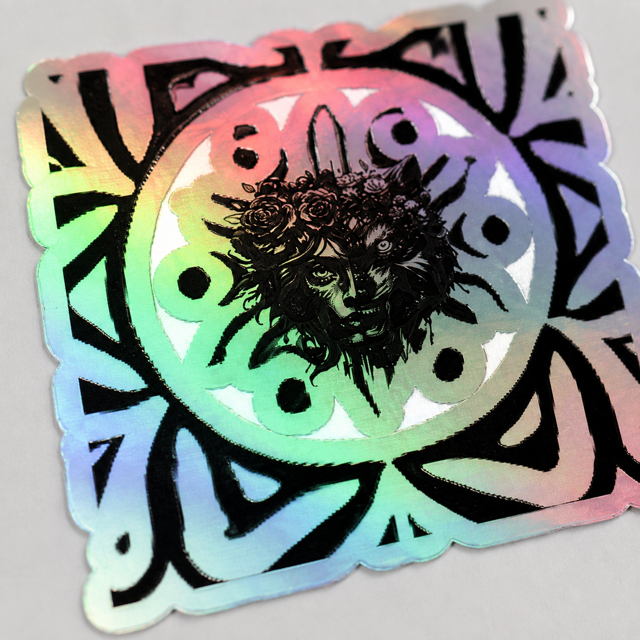 A square holographic sticker with a black sun-and-moon style graphic and a central dark face illustration, photographed on a light gray background.