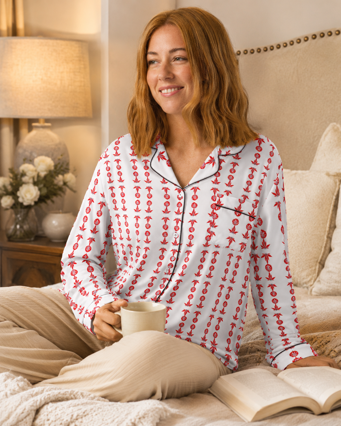 A woman sitting on a bed wearing a white long-sleeve pajama shirt with a red anchor pattern and beige pants, holding a mug and reading a book in a cozy bedroom setting.
