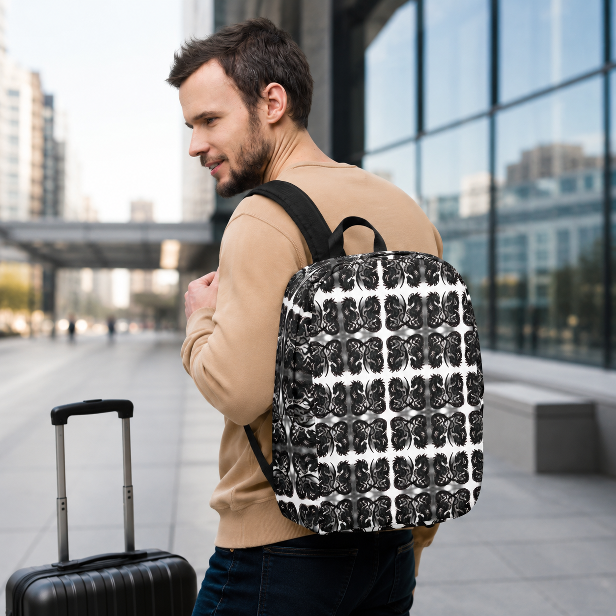 A man wearing a beige sweater and dark jeans carries a black and white patterned backpack while standing in a modern outdoor urban setting with a suitcase nearby.
