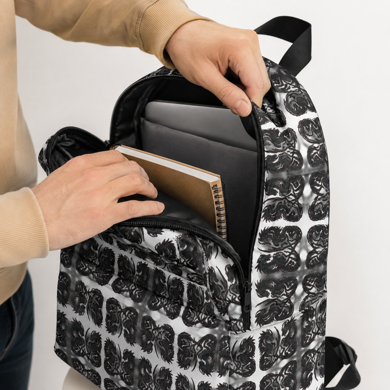 A close-up of a backpack with a black and white skull pattern, shown open with a laptop and notebook inside. A person's hands and arms are visible interacting with the bag against a plain white background.