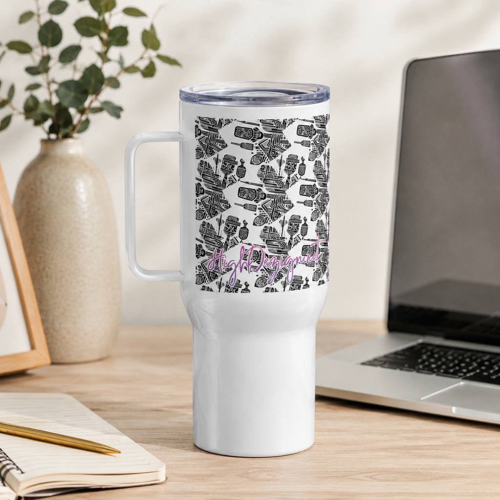 A white travel tumbler with a black floral pattern and purple script text, shown on a wooden desk next to a laptop, a vase with greenery, and an open notebook.
