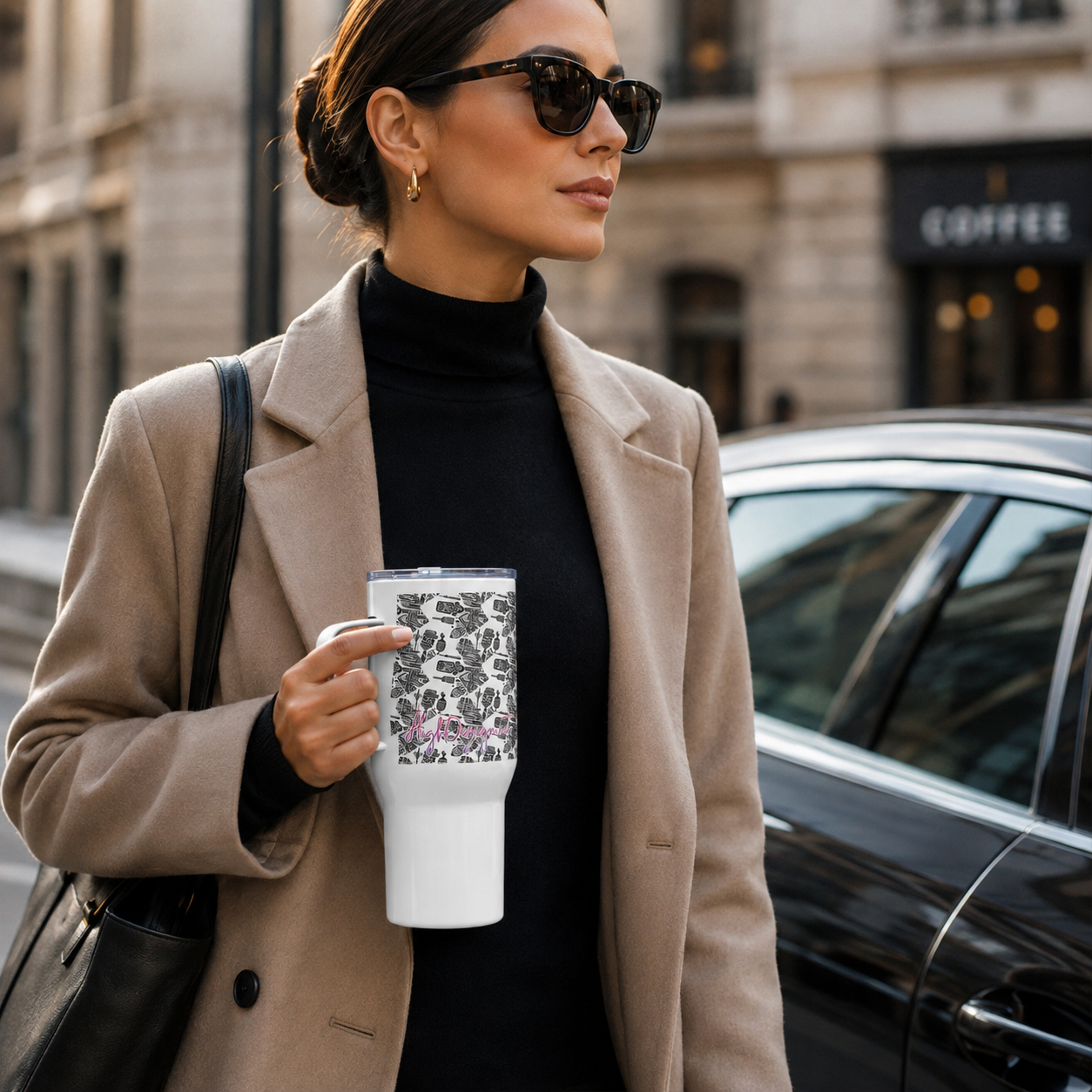 A woman in a beige blazer and black turtleneck holds a white insulated tumbler with a black floral pattern and pink script text on a city street next to a black car.