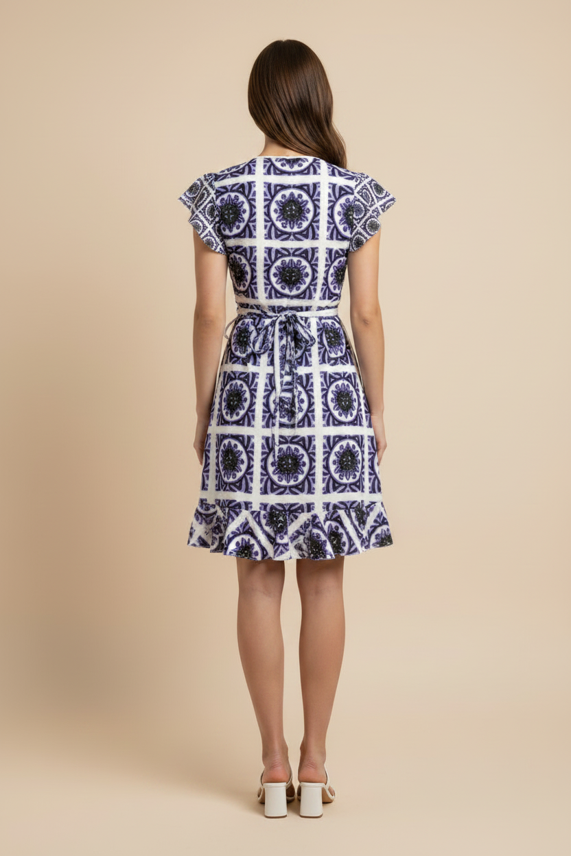 A woman modeling a short V-neck dress with a blue and white geometric pattern, ruffled cap sleeves, and a flared skirt, standing against a solid beige background.