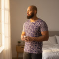 Man in a floral shirt standing in a bedroom holding a mug.