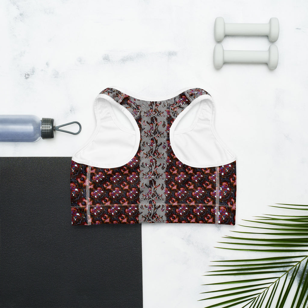 A women's sports bra with a red, black, and gray patterned design and white underband, styled in a flat lay on a white marble background with a black yoga mat, dumbbells, a water bottle, and a palm leaf.