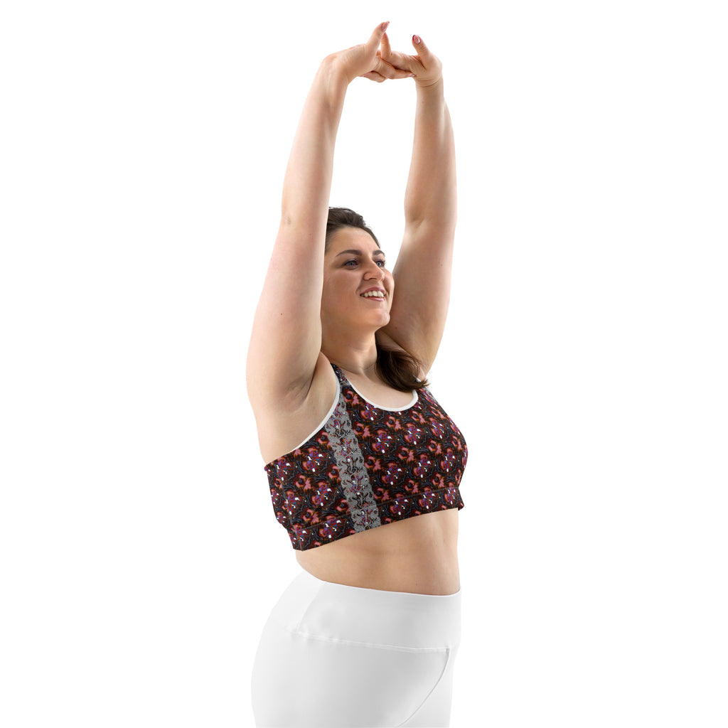 A woman wearing a floral-patterned sports bra and white leggings poses with her arms raised overhead against a solid white background.